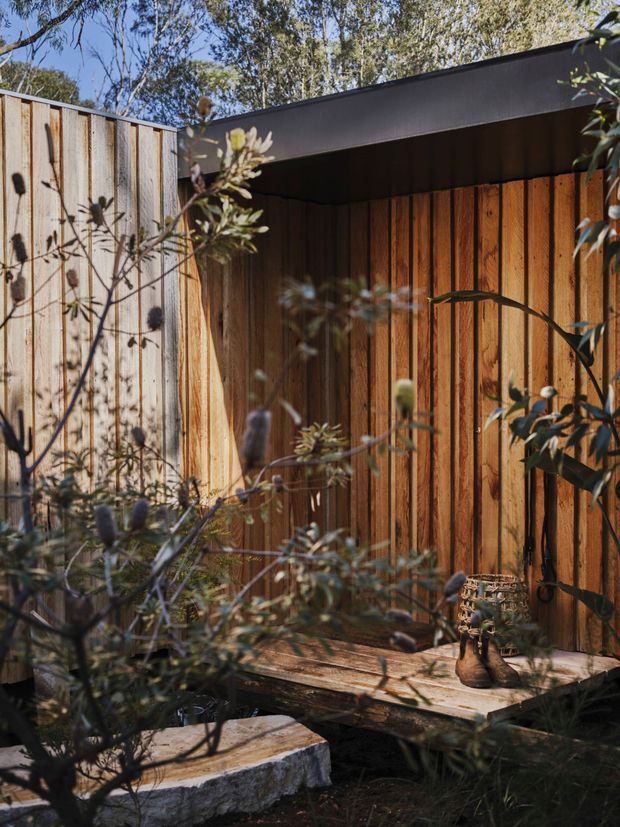 A secluded outdoor courtyard or garden corner enclosed by vertical timber fencing and cladding. A dark, flat-roofed canopy creates a sheltered alcove over a rustic timber bench or deck, with natural stone edging and dense native planting in the foreground. The warm weathered wood, black roof trim, and dappled sunlight create an intimate, organic outdoor setting.