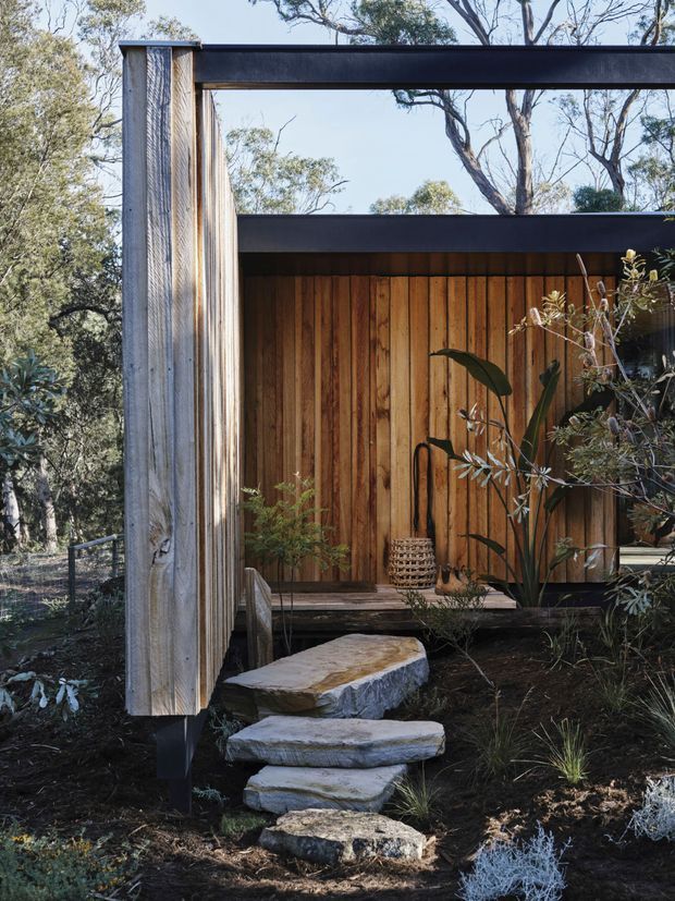 A close exterior view of a compact timber-clad structure set within a natural bush landscape. Vertical weathered timber boards form the walls, framed by dark horizontal steel or painted metal beams and a projecting flat roof. Large irregular stone stepping slabs lead through planted garden beds to a small raised entry deck. Native vegetation and mature trees surround the structure, creating a restrained, organic architectural composition.
