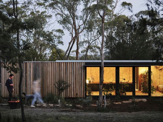 A low, contemporary single-storey dwelling set within a wooded landscape. The facade combines vertical natural timber cladding with a dark flat roof and extensive floor-to-ceiling glazing, revealing a warmly lit interior. A landscaped garden and mature trees surround the building, while blurred figures provide a sense of scale.