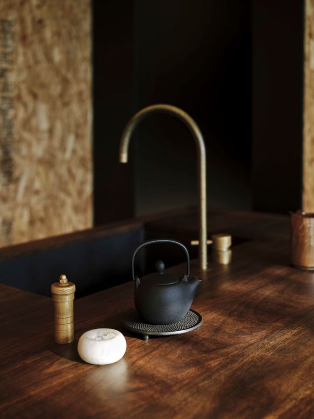 Close-up of a dark, richly toned kitchen benchtop featuring a matte black kettle on a trivet, a tall brushed brass gooseneck tap, a wooden pepper mill, and a small white vessel. Dark cabinetry or wall surfaces and a textured timber-toned wall create a moody, refined kitchen vignette.