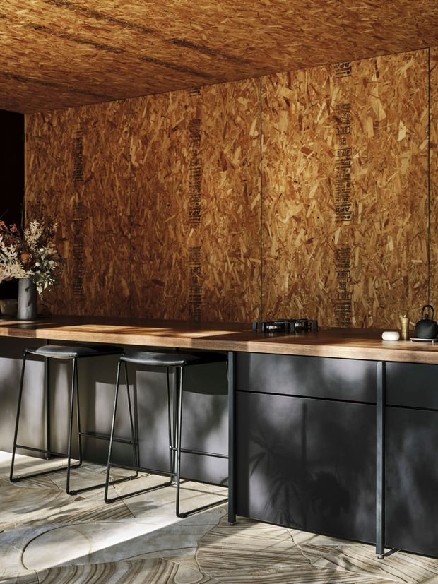 A compact, moody kitchen with exposed oriented strand board (OSB) walls and ceiling, a long timber benchtop, and matte black built-in cabinetry. A black cooktop, kettle, and small accessories sit on the counter, with three slim black bar stools along the island or peninsula. Patterned stone-look flooring and strong natural shadows add texture and contrast.