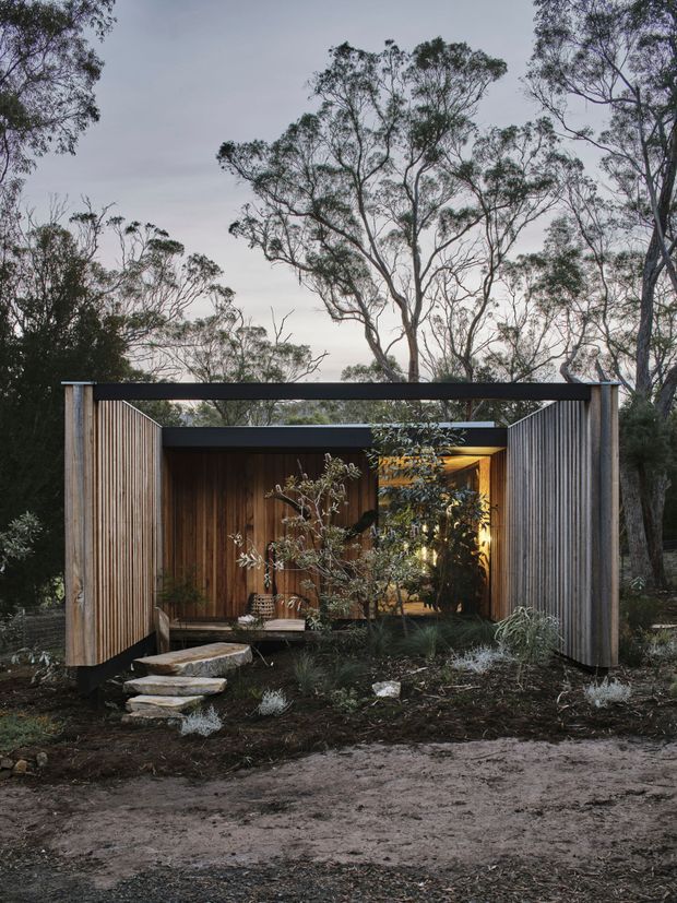 A compact modern pavilion or dwelling set within a native woodland landscape. The structure features a dark flat roof, warm vertical timber cladding, and large vertically slatted timber screens forming a framed entry. A simple stone stepping path leads through a low, naturalistic garden toward the warmly illuminated interior. The composition has a quiet, immersive connection to the surrounding trees and appears photographed at dusk.