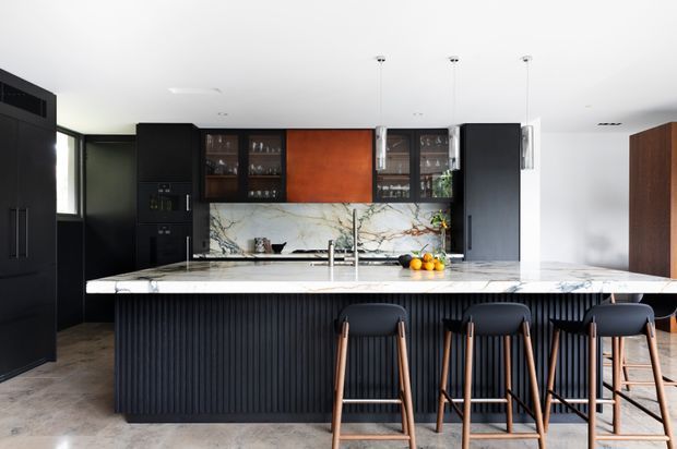 A contemporary kitchen viewed straight on, featuring a large marble island with a dark vertically fluted base, black cabinetry, glazed display cupboards, a warm timber-toned central cabinet panel, and a marble-look veined splashback. Three pendant lights hang above the island, with bar stools arranged along the front.
