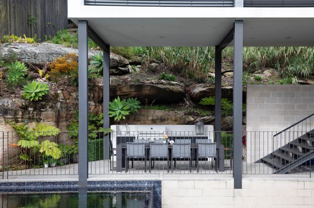 Contemporary elevated outdoor dining area beside a pool, with a dark metal-framed upper structure, concrete block wall, open stairway, and a natural rocky landscaped embankment behind the dining setting.