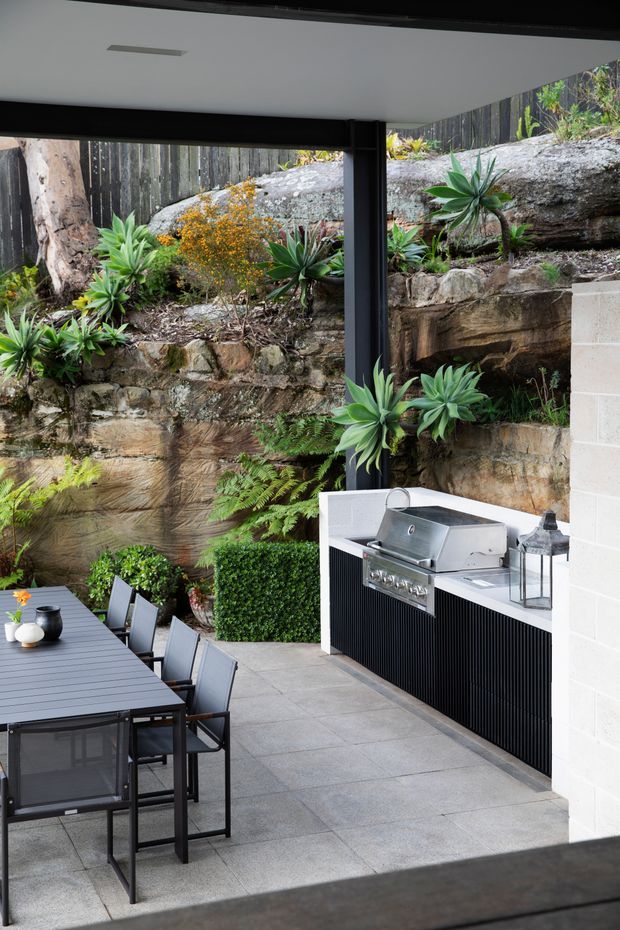 A covered outdoor entertaining area with a built-in stainless-steel barbecue, black ribbed cabinetry, dining table and chairs, and large-format grey paving. The space opens onto a landscaped, terraced garden with exposed rock retaining walls, layered greenery and timber fencing. Black structural framing and a white ceiling create a sheltered, contemporary outdoor kitchen and dining setting.