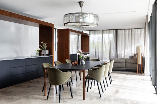 A refined open-plan dining area with a long dark dining table surrounded by upholstered olive-grey chairs. Floor-to-ceiling sheer curtains provide abundant natural light, while dark timber-framed wall panels and built-in cabinetry contrast with pale stone-look flooring. A large crystal-style circular pendant light hangs above the table, and a lounge area is visible beyond.