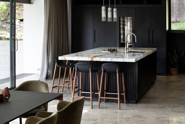A sophisticated open-plan kitchen and dining area with a large dark island, dramatic veined stone benchtop, integrated black cabinetry, pendant lights, and timber-framed bar stools. A dining table and upholstered chairs are visible in the foreground, with full-height glazing and curtains along the left side.