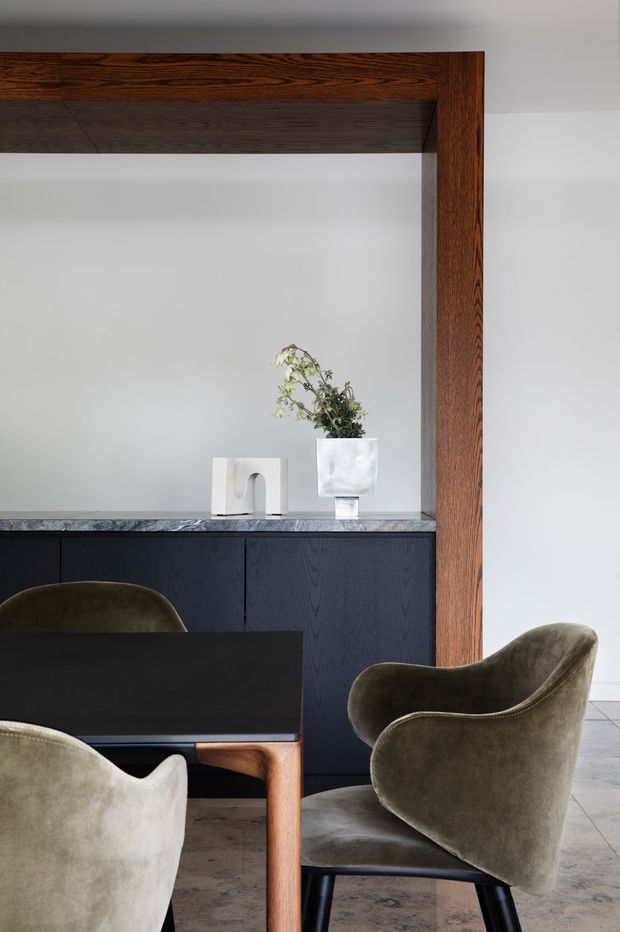 A refined dining area with a dark rectangular dining table and sculptural upholstered dining chairs in muted olive-grey tones. A dark navy-blue built-in cabinet runs along the rear wall beneath a grey stone countertop, with minimalist objects and a vase displayed above. Strong vertical and horizontal dark timber framing surrounds the wall opening, contrasting with pale plaster walls and a large-format neutral tiled floor.