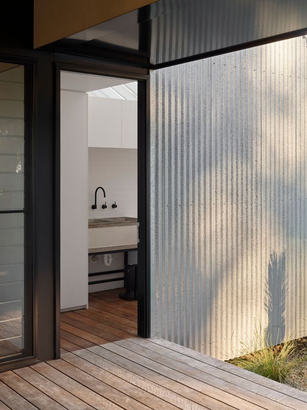 A modern exterior threshold showing a timber deck and dark-framed glazed doorway opening into a compact bathroom or washroom. The interior has white wall surfaces, a concrete-look vanity benchtop with black wall-mounted tapware, exposed plumbing beneath the basin, and timber flooring. A large translucent corrugated-panel wall admits filtered daylight, creating a strong vertical texture alongside the deck.