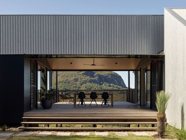 Exterior view of a contemporary, pavilion-like home with a covered timber deck and outdoor dining area. Large glazed openings frame a mountainous landscape, while dark corrugated cladding, pale textured wall cladding, timber decking and steps, and a simple pitched roof create a restrained modern composition.