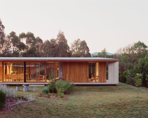 Single-storey contemporary residential pavilion set within a landscaped garden, with extensive floor-to-ceiling glazing, warm vertical timber cladding, a low-profile corrugated metal roof, and a contrasting pale textured end wall. The elevated structure opens onto a grassy garden and reveals a warm timber-lined interior with dining and living areas.