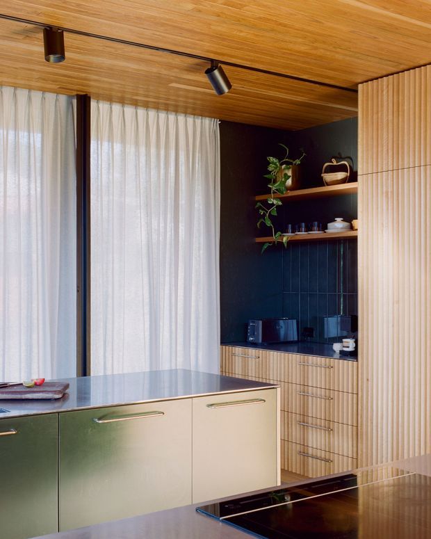 A refined contemporary kitchen with a central island, pale cabinetry, dark stone benchtops, vertical fluted timber joinery, open shelving, and a dark tiled backsplash. The room features a warm timber-lined ceiling with black track lighting, sheer curtains across large glazing, integrated appliances, and subtle greenery and ceramics on the shelves.