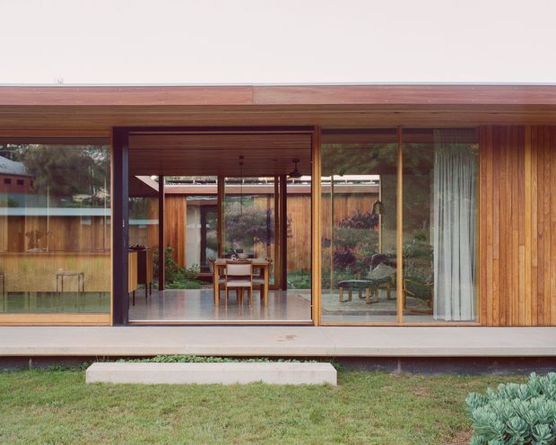 A low, single-storey modern dwelling viewed from the garden, with extensive floor-to-ceiling glazing, timber-framed sliding doors, warm vertical timber cladding, and a broad shallow roof overhang. The open glazing reveals a dining area and a planted courtyard or garden beyond, creating a strong indoor–outdoor connection. The composition is calm and rectilinear, with dark structural framing and restrained landscaping.