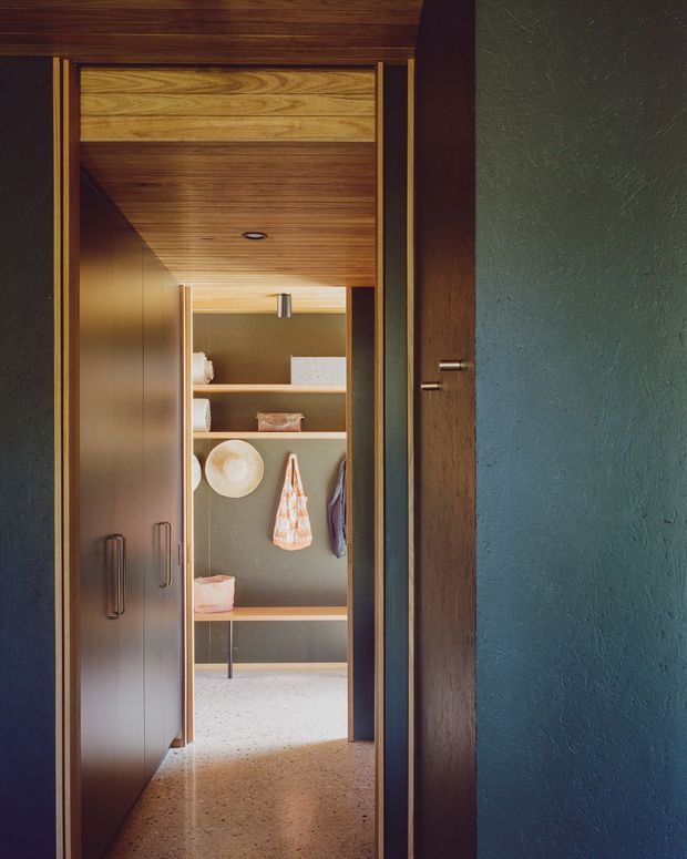 View through a dark timber-framed doorway into a compact, carefully fitted storage and dressing area. The space features full-height dark cabinetry, open timber shelving, hanging garments and accessories, a warm timber-lined ceiling, muted green textured walls, and a polished terrazzo floor. The composition emphasises the layered doorway, built-in joinery, and warm natural materials.