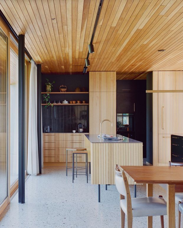 A refined open-plan kitchen and dining area with extensive vertically fluted timber cabinetry, a matching timber-lined ceiling, dark wall finishes, and a central island with an integrated sink. The room has full-height glazing along one side, a pale terrazzo-like floor, track lighting, open shelving, and a warm timber dining table with upholstered chairs.