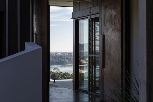 A dark, refined interior threshold opens onto a timber deck with expansive black-framed glazing and a scenic view of a waterway and distant hills. Vertical dark timber wall cladding, a large textured timber pivot door, and a suspended chain detail create a dramatic contemporary architectural setting.