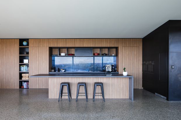Contemporary open-plan kitchen dominated by a long timber-lined wall of integrated cabinetry and shelving, a central island with a dark benchtop and three stools, and a large blue-toned splashback or window behind the cooktop. The space has a polished speckled concrete or terrazzo floor, dark cabinetry or wall joinery at right, and a restrained, refined palette of timber, charcoal, grey, and blue.