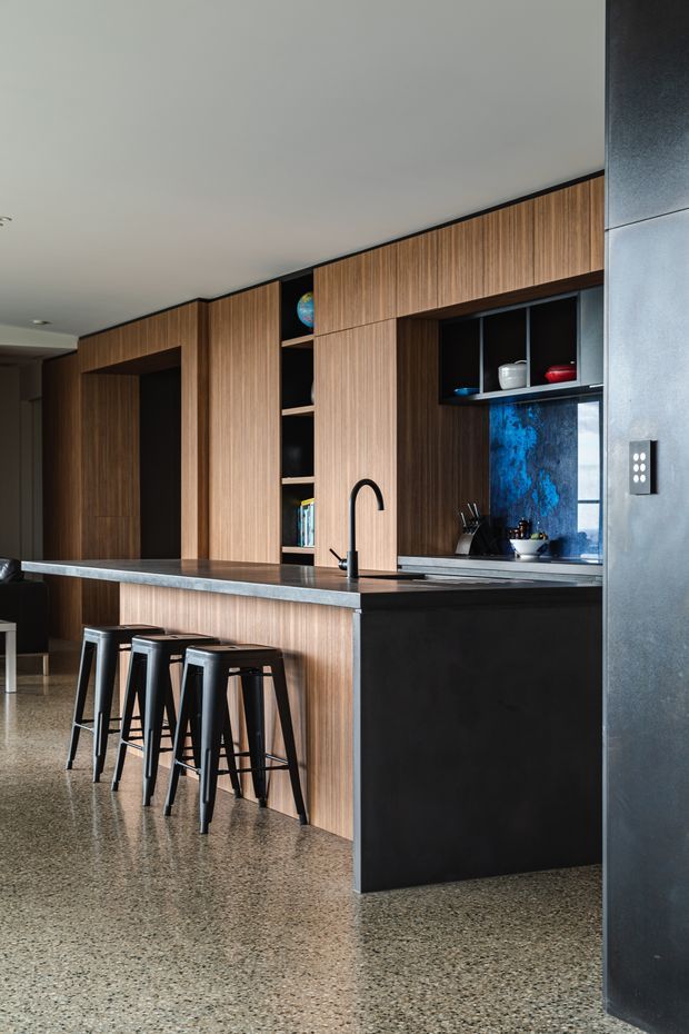 A contemporary kitchen with a long island and integrated sink, finished in dark timber veneer cabinetry with black panels and shelving. The island has a grey stone benchtop and three black bar stools. A blue textured splashback adds colour behind the cooktop area, while a terrazzo floor and dark feature wall create a refined, moody scheme.
