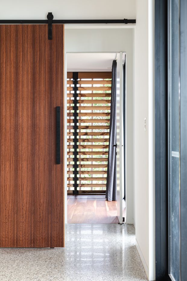 Interior view of a contemporary entryway or hallway featuring a large dark timber sliding barn-style door with black metal track and handle. The polished terrazzo floor leads toward an adjoining space with warm timber flooring, horizontal timber screening, and a dark curtain. White walls and dark-framed glazing create a bright, refined transition between spaces.