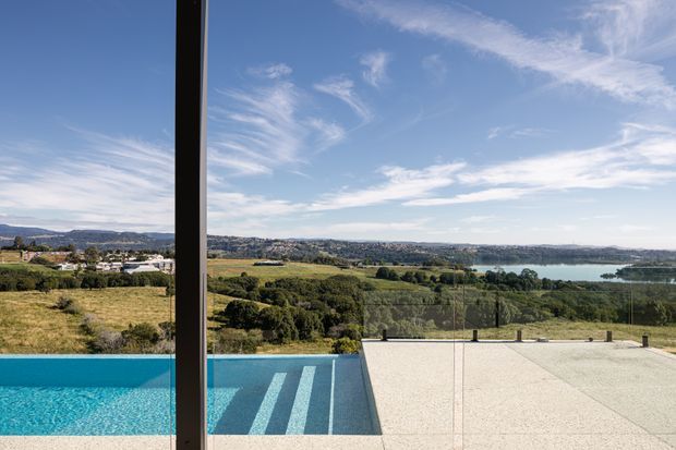 A wide architectural view from a contemporary property overlooking a rural, hilly landscape and a broad body of water. The foreground features a turquoise swimming pool, pale paved terrace, and a frameless glass balustrade, with a dark vertical window or door frame dividing the view. The scene is bright, open, and strongly connected to the surrounding landscape.