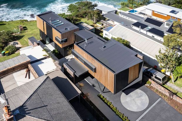 Aerial view of a contemporary multi-building residential development on a coastal site. Several dark metal-roofed buildings feature vertical timber cladding, large roof skylights, and modern rectangular forms, connected by paved driveways and paths. The ocean and surf are visible directly beyond the property.