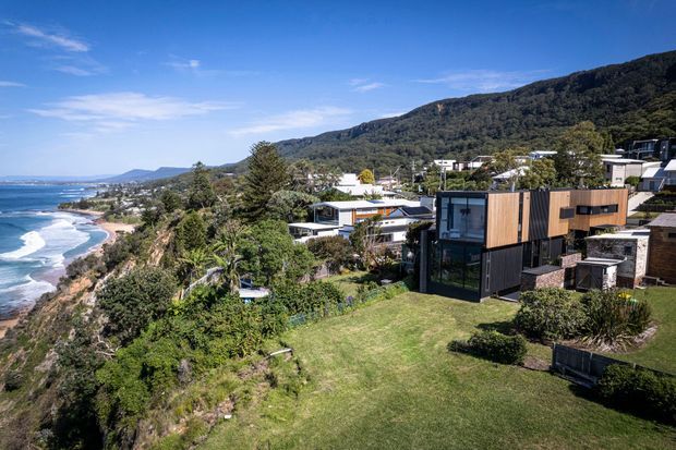 Aerial view of a contemporary coastal residential neighbourhood on a steep, landscaped site, with a modern dark-clad and timber-clad house in the foreground. The homes overlook a beach and ocean, backed by a densely forested mountain ridge under a clear blue sky.