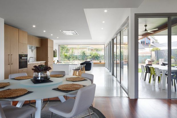 Bright open-plan contemporary kitchen and dining area with extensive glazing. A pale dining table with upholstered chairs occupies the foreground, while timber kitchen cabinetry, integrated appliances and a stone island are visible to the left. Full-height sliding glass doors open to an outdoor dining area, and polished light flooring transitions to darker timber flooring in the dining zone.
