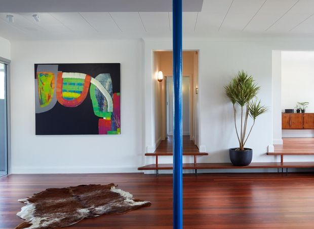 Bright, open-plan mid-century-inspired interior featuring polished dark timber flooring, white walls and a white paneled ceiling. A vivid abstract artwork is mounted on the left wall, while a long built-in timber bench or low console runs across the room. A vivid blue structural or decorative vertical pole divides the composition, with a potted indoor plant and a cowhide rug adding natural texture. An open hallway and timber sideboard are visible in the background.