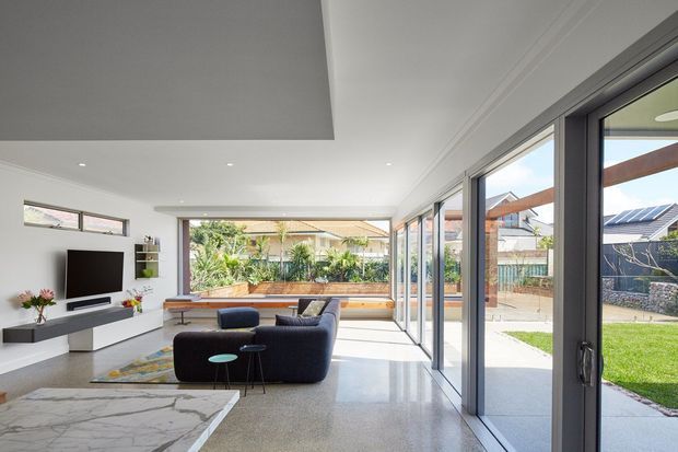 Bright contemporary open-plan living room with a low-profile media wall, dark modular seating, polished light-toned flooring, and extensive floor-to-ceiling glazing opening to a landscaped backyard. A marble-look surface is visible in the foreground, while timber-framed outdoor elements and neighbouring homes can be seen through the glazing.