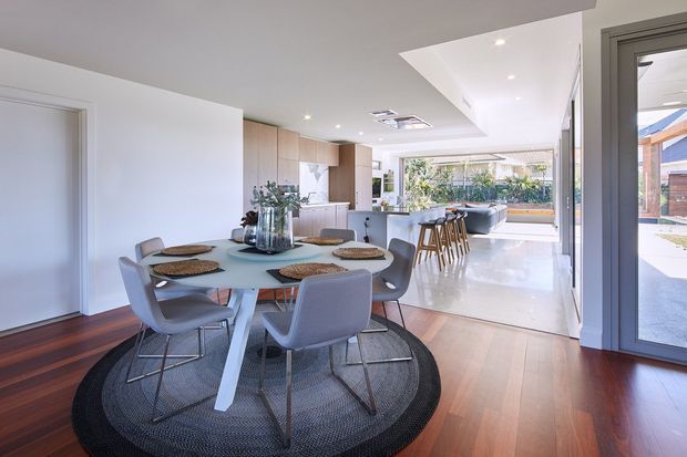 Open-plan dining and kitchen area in a bright contemporary home. A round dining table with upholstered chairs sits on a large circular rug over dark timber flooring, while the kitchen beyond features light wood cabinetry, a pale island benchtop, and integrated indoor-outdoor glazing opening toward a landscaped courtyard.