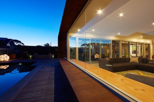 Twilight exterior view of a contemporary home, looking along a broad timber-framed glazed wall into a warmly lit living room. A dark timber deck and reflecting swimming pool occupy the foreground, with extensive floor-to-ceiling glazing connecting the interior to the outdoor area.