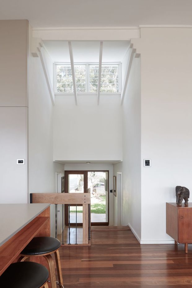 Bright, narrow double-height interior circulation space with a timber staircase and glass-panel entry door at the lower level. White walls and ceiling are accented by exposed white ceiling beams and clerestory-style glazing that brings in abundant daylight. A timber kitchen or built-in counter with black stools is visible in the foreground, while a mid-century-style timber sideboard sits to the right.
