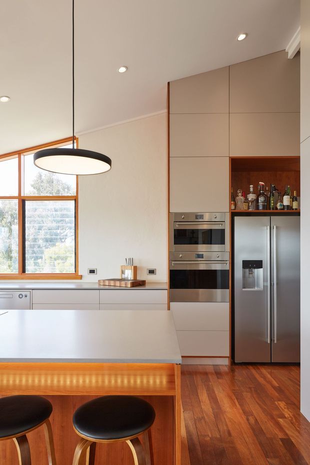 A bright contemporary kitchen with a large pale island and integrated timber breakfast-bar extension, paired with two black round stools. The room features full-height warm taupe cabinetry, stainless-steel double ovens and a large stainless-steel French-door refrigerator, plus an open timber-lined drinks display niche. Timber-framed windows bring in natural light, while recessed ceiling lights and a large black-and-white pendant provide layered illumination. Warm timber flooring and cabinetry accents create a refined, inviting atmosphere.