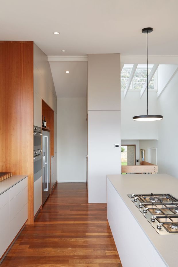 Bright contemporary kitchen interior with a long white island and integrated gas cooktop, full-height warm timber cabinetry, white built-in appliances, polished timber flooring, and a high ceiling with skylights. A pendant light hangs above the island, while an open connection to a lower living area is visible beyond.
