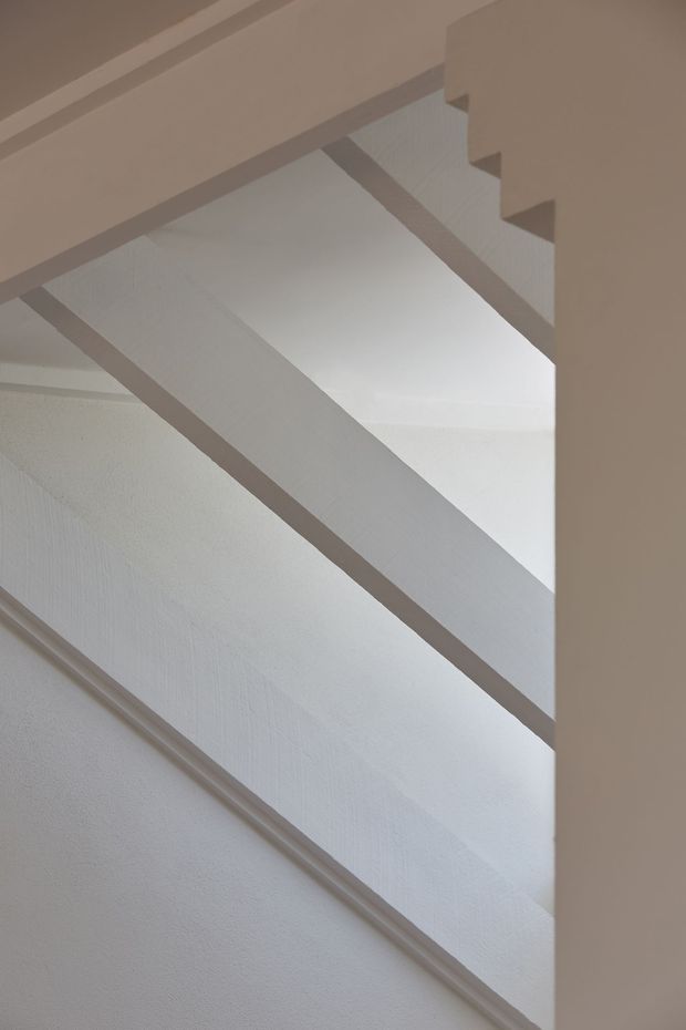 Close-up architectural view of a bright white stairway with diagonal stair stringers, textured painted walls, simple trim, and the underside of a staircase. The composition is minimalist and focused on geometric lines and natural-looking daylight.