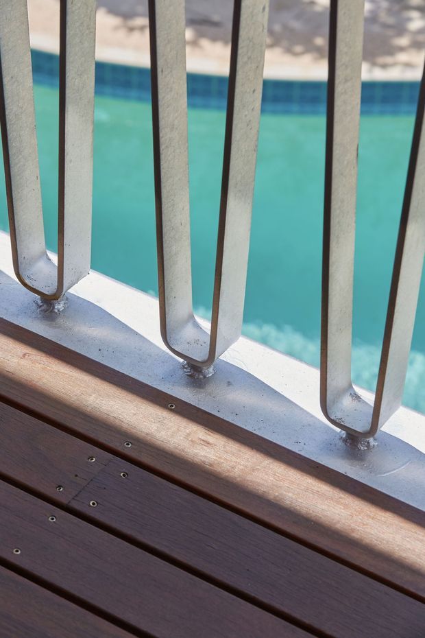Close-up architectural detail of a timber deck or balcony floor beside a swimming pool, with a painted metal balustrade featuring repeated U-shaped vertical rails. Bright turquoise pool water is visible beyond the railing.