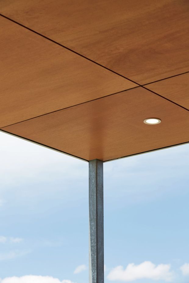 Close-up architectural view from beneath a projecting timber-lined roof or canopy, showing clean panel joints, a recessed round downlight, and a slender exposed steel support column against an open sky background.