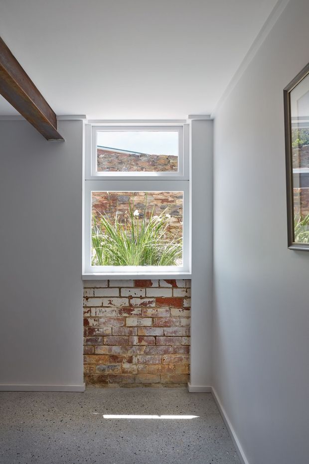 A narrow, bright interior space with white painted walls, a white-framed two-part window, exposed reclaimed brickwork beneath the window, and a polished speckled concrete or terrazzo-like floor. A dark timber beam or shelf runs along the upper left wall, while a framed reflective surface is visible on the right. The window looks onto a brick boundary wall and ornamental grasses, creating a clean industrial-heritage character.