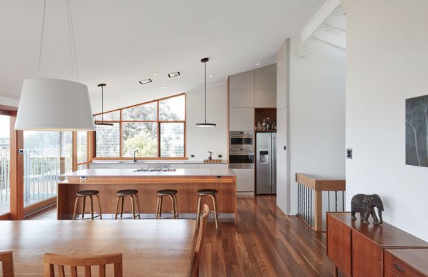 Open-plan kitchen and dining area with a timber island, four bar stools, integrated stainless-steel appliances, warm timber flooring, and large timber-framed windows. A dining table and mid-century-style sideboard occupy the foreground, while a stair opening and white structural ceiling details are visible to the right. The room is bright and warm with a clean contemporary fit-out.