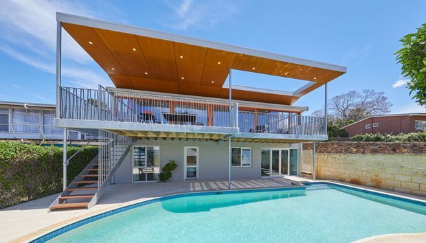 Exterior view of a contemporary multi-level residence with a large turquoise swimming pool in the foreground. The house features elevated decks and balconies with metal balustrades, an open steel-framed structure, broad flat projecting roof canopies with warm timber soffits and recessed downlights, extensive glazing, and an external stair. A rendered lower level, masonry boundary wall, and landscaped surroundings complete the scene.