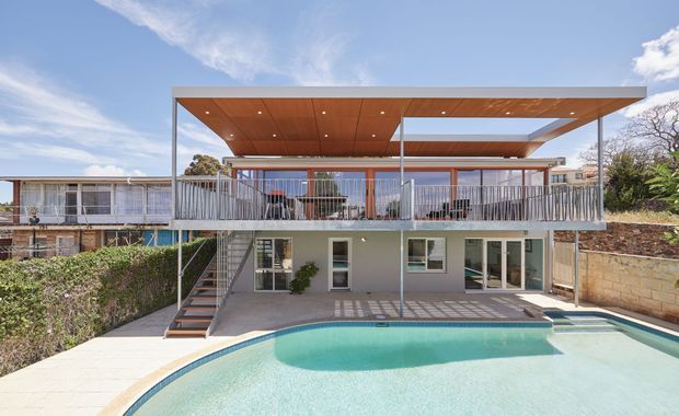 Rear exterior view of a contemporary two-level home with a large covered upper terrace, timber-lined soffit, slim metal posts and balustrades, external stairway, ground-level glazing, paved pool surround, and a swimming pool in the foreground. The elevated terrace and open roof canopy are the dominant architectural features.