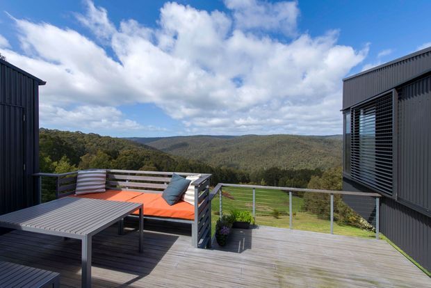 A modern elevated outdoor terrace between dark corrugated-metal building volumes, overlooking a densely wooded rolling landscape. The terrace has grey timber decking, a metal-framed dining table and bench seating with bright orange and blue cushions, and a simple horizontal railing. The scene is bright and expansive beneath a partly cloudy blue sky.