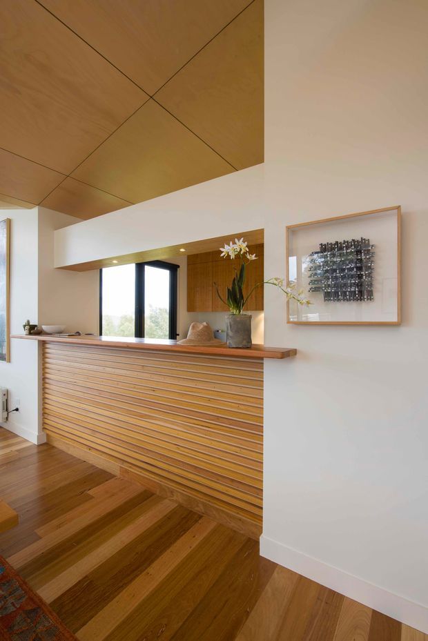A warm, refined residential interior showing a timber-lined stairway and landing beside a built-in slatted timber balustrade or screen. The space features rich timber flooring, a timber ceiling with geometric panel joints, white plaster walls, dark-framed floor-to-ceiling glazing, recessed lighting, and a timber ledge displaying a potted flowering plant. A framed sculptural artwork hangs on the wall.