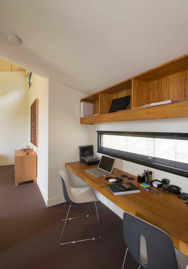 Compact home office with a long timber desk beneath a wide horizontal window, open timber shelving and overhead storage, two modern molded chairs, and office equipment. The room has white walls and ceiling, dark carpet, warm wood joinery, and abundant daylight.