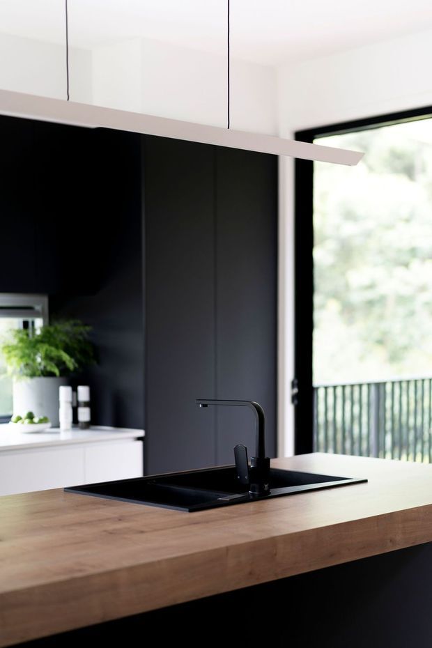 Close-up view of a contemporary kitchen island with a thick natural timber benchtop, integrated black sink and matte black gooseneck tap. Dark full-height cabinetry contrasts with white overhead cabinetry and walls. A large black-framed glazed opening provides views to greenery and a railing outside.