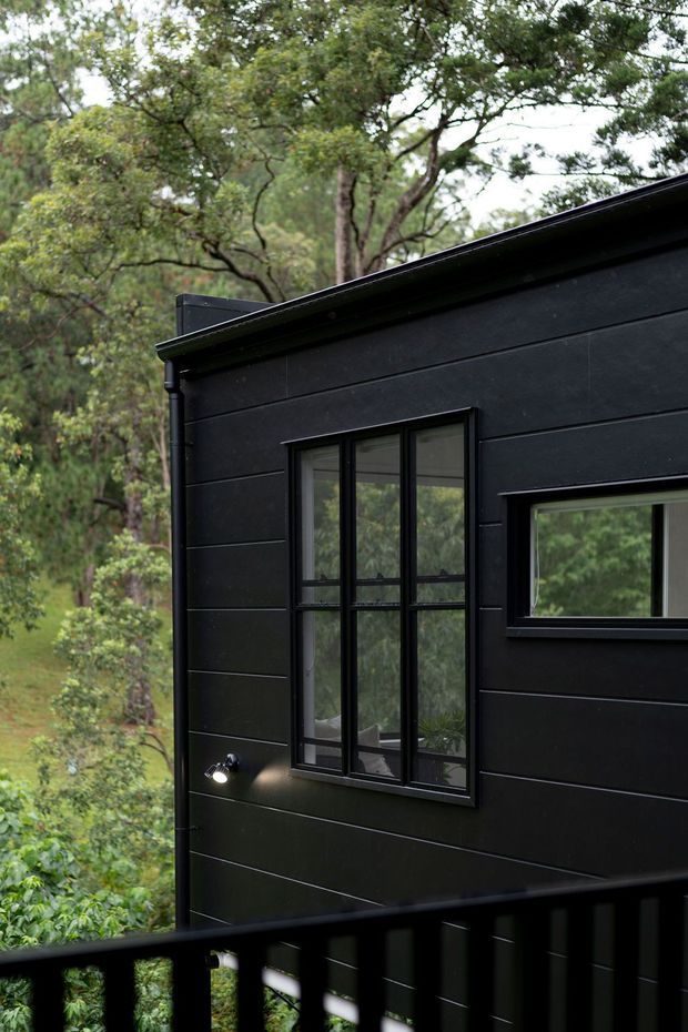 Exterior view of a compact dark-clad building set among dense trees. The facade has horizontal black cladding, black-framed vertical and horizontal windows, a simple roof edge with guttering, an exterior wall light, and a black deck or balcony railing in the foreground.