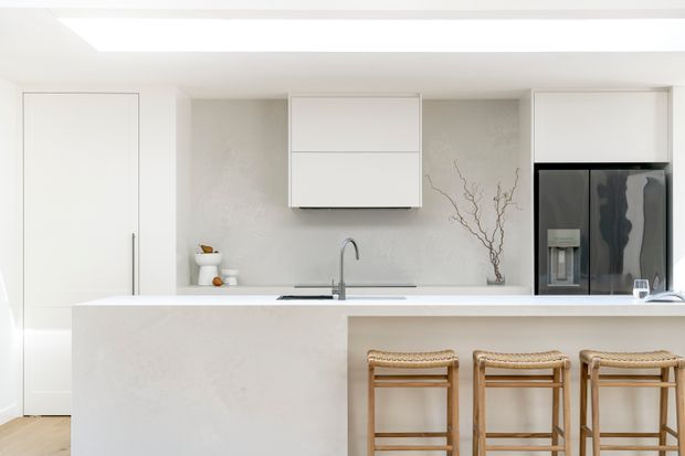 Bright, minimalist contemporary kitchen with a large pale stone island, integrated white cabinetry, a concealed tall pantry or appliance cabinet, pale textured backsplash, undermount sink, black refrigerator, and three woven-seat timber bar stools. The space has clean lines, restrained neutral colours, and abundant daylight.