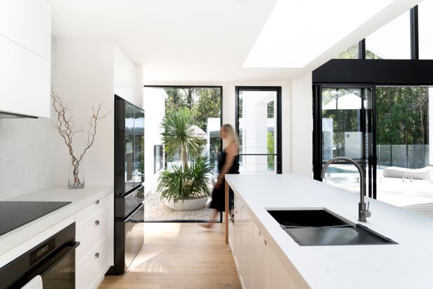 Bright contemporary kitchen with white flat-panel cabinetry, a large white island with integrated black sink and curved chrome tap, pale timber flooring, and expansive black-framed glazing. The open-plan space looks onto a landscaped courtyard and includes a large skylight and abundant natural light.