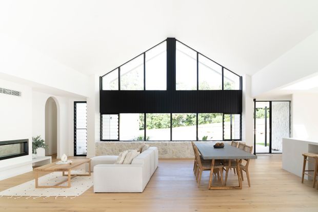 Bright open-plan living and dining area with white walls, pale timber flooring, a large double-height black-framed glazed feature wall, exposed stone fireplace surround, white sectional sofa, timber coffee table, and timber dining table. The space opens toward a kitchen area and an exterior courtyard or garden.
