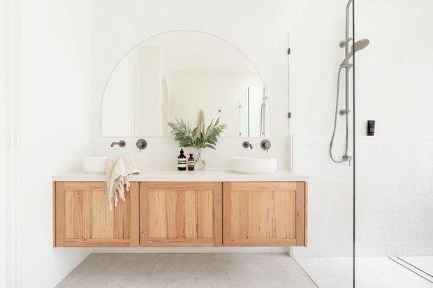 Bright, minimalist bathroom featuring a wall-mounted natural timber double vanity with two white basins, a large arched mirror, wall-mounted fixtures, and a frameless glass walk-in shower. White tiled walls and a pale floor create a clean, airy aesthetic.